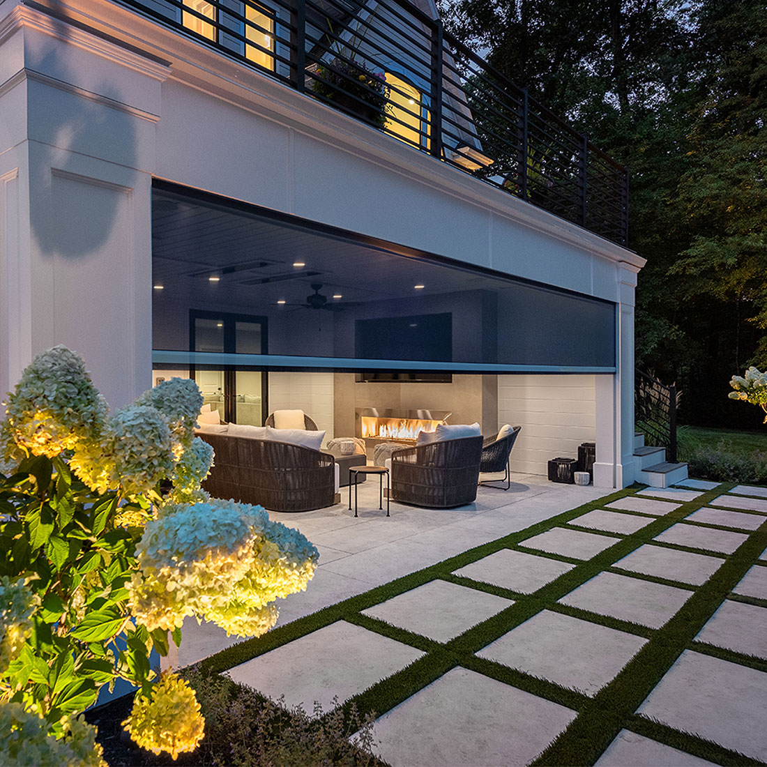 Phantom motorized wall screen partially lowered across a covered outdoor living space with a seating area and fireplace at dusk